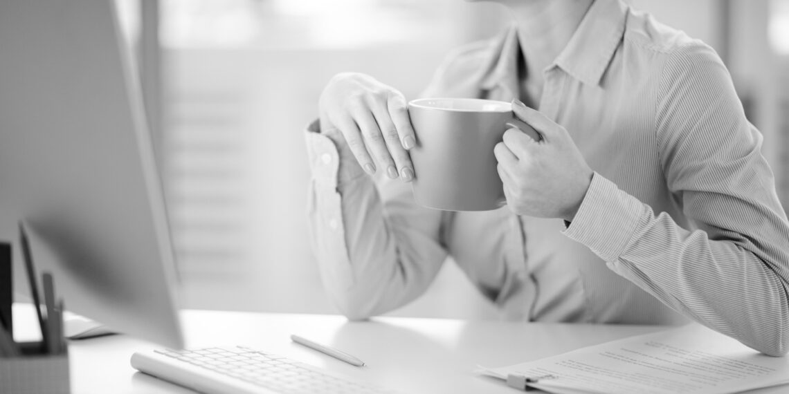 Young woman with yellow mug sitting by desk in office in front of computer monitor